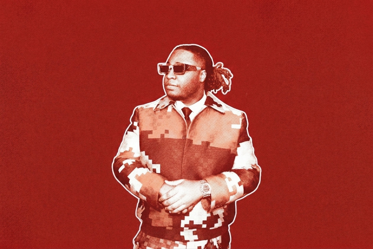 Tay Keith wearing a camouflage-patterned jacket and sunglasses stands in front of a Grammy-themed backdrop.