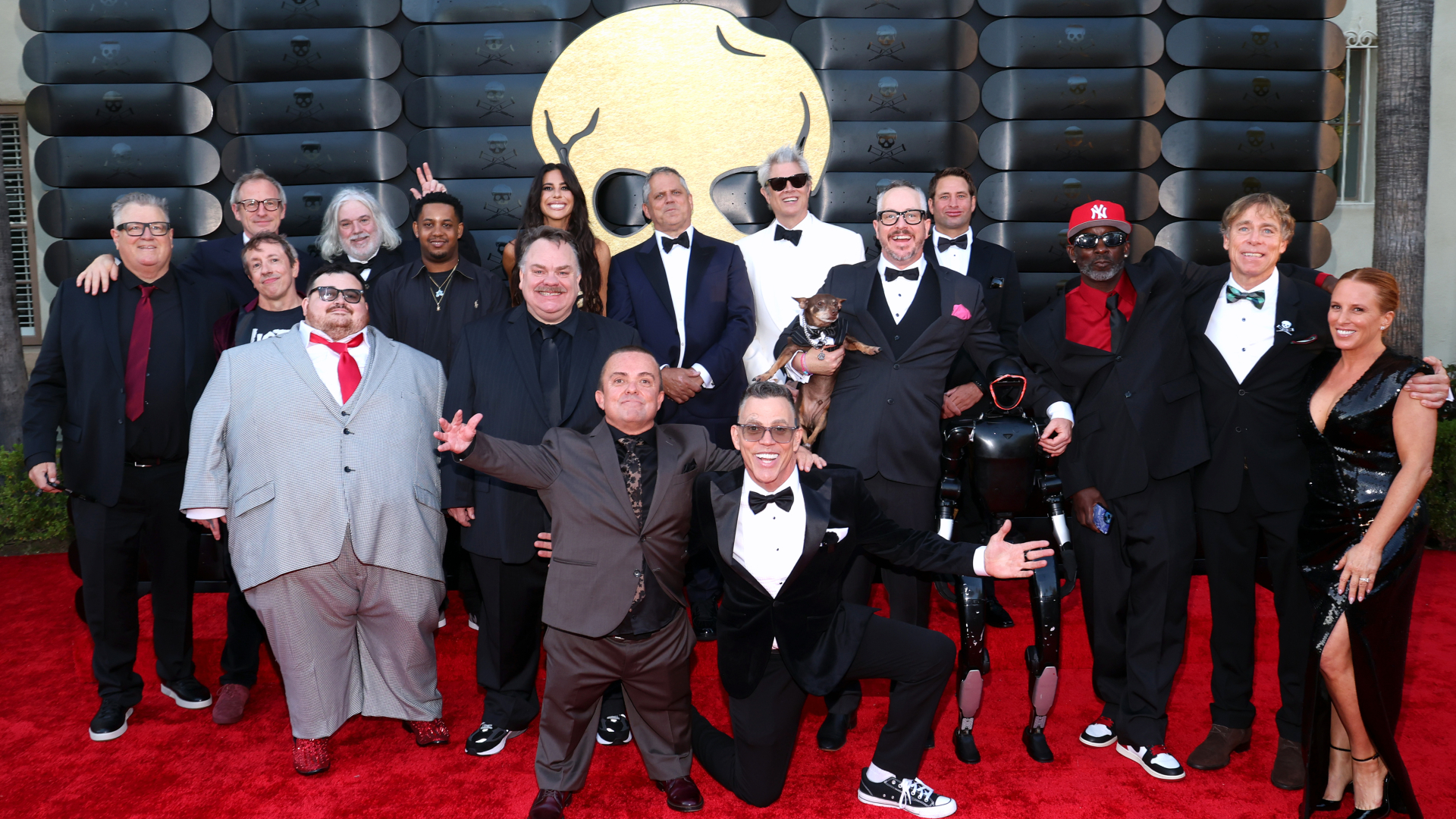 A group of stars, including the cast of "Jackass," pose on a red carpet in formal attire with a large logo in the background.