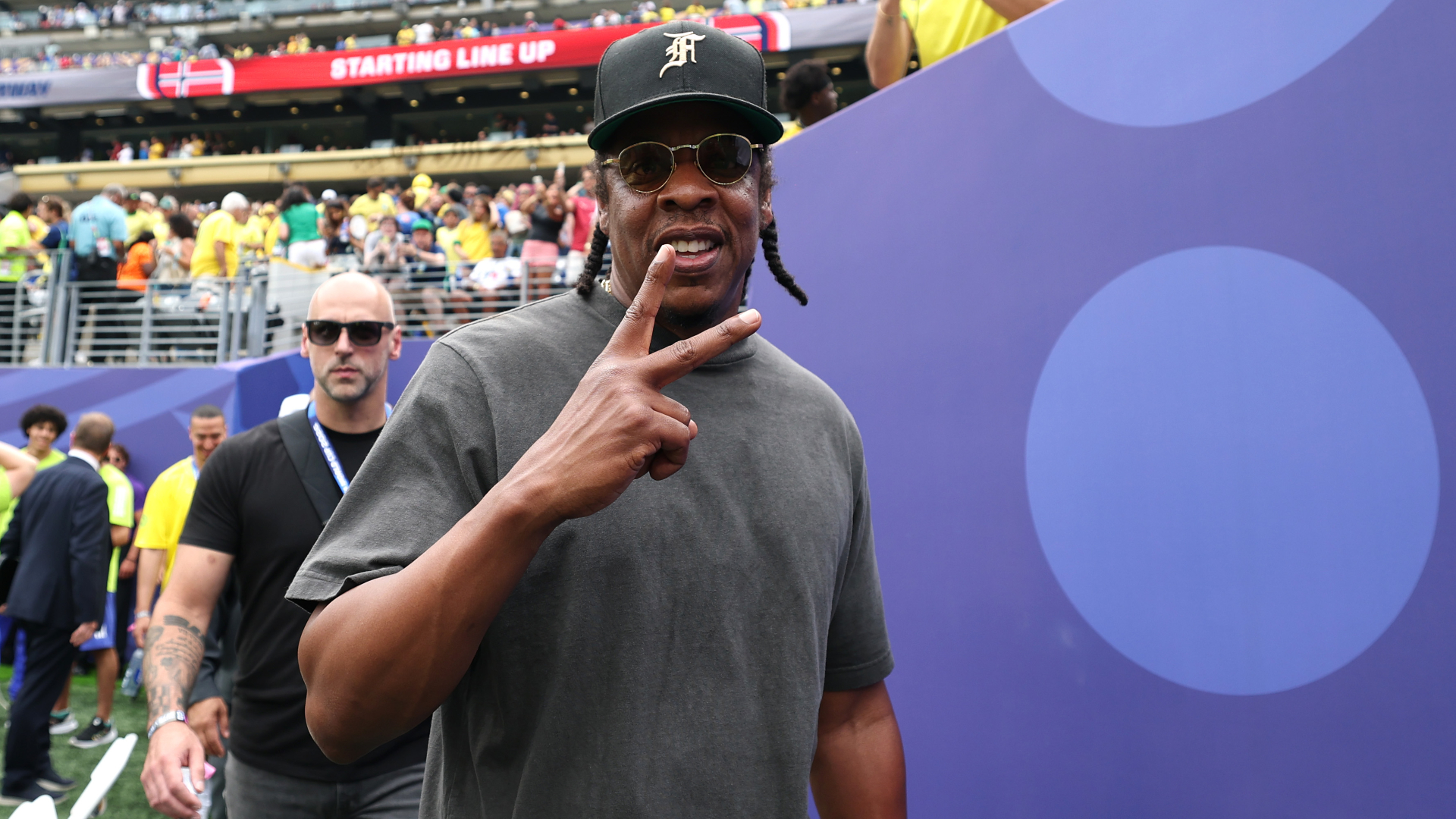 Jay-Z wearing sunglasses and a cap, making a peace sign at a sports event.