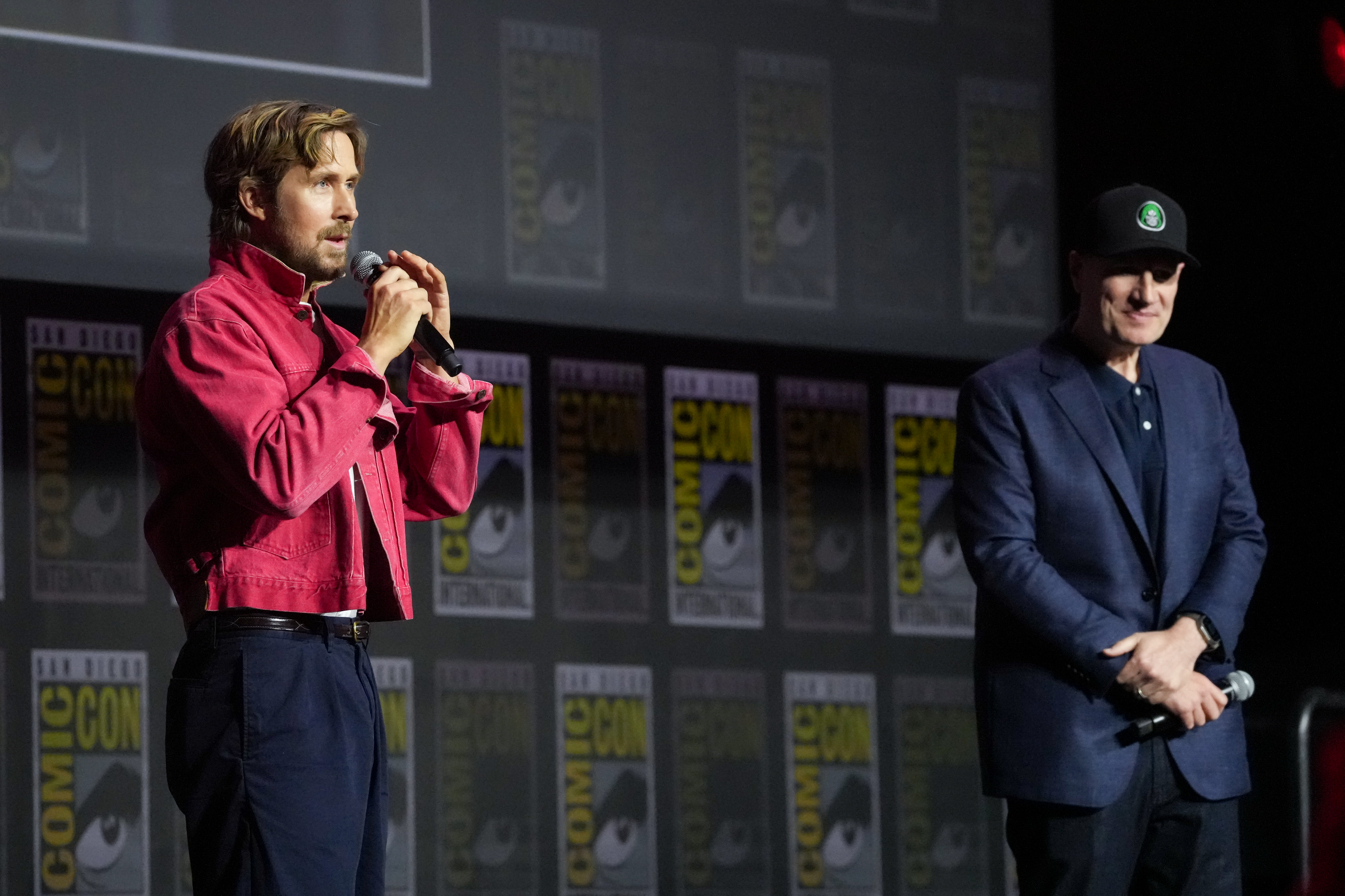 Ryan Gosling in a red jacket speaks into a microphone on stage, while Kevin Feige in a cap and blazer stands beside him at Comic-Con.