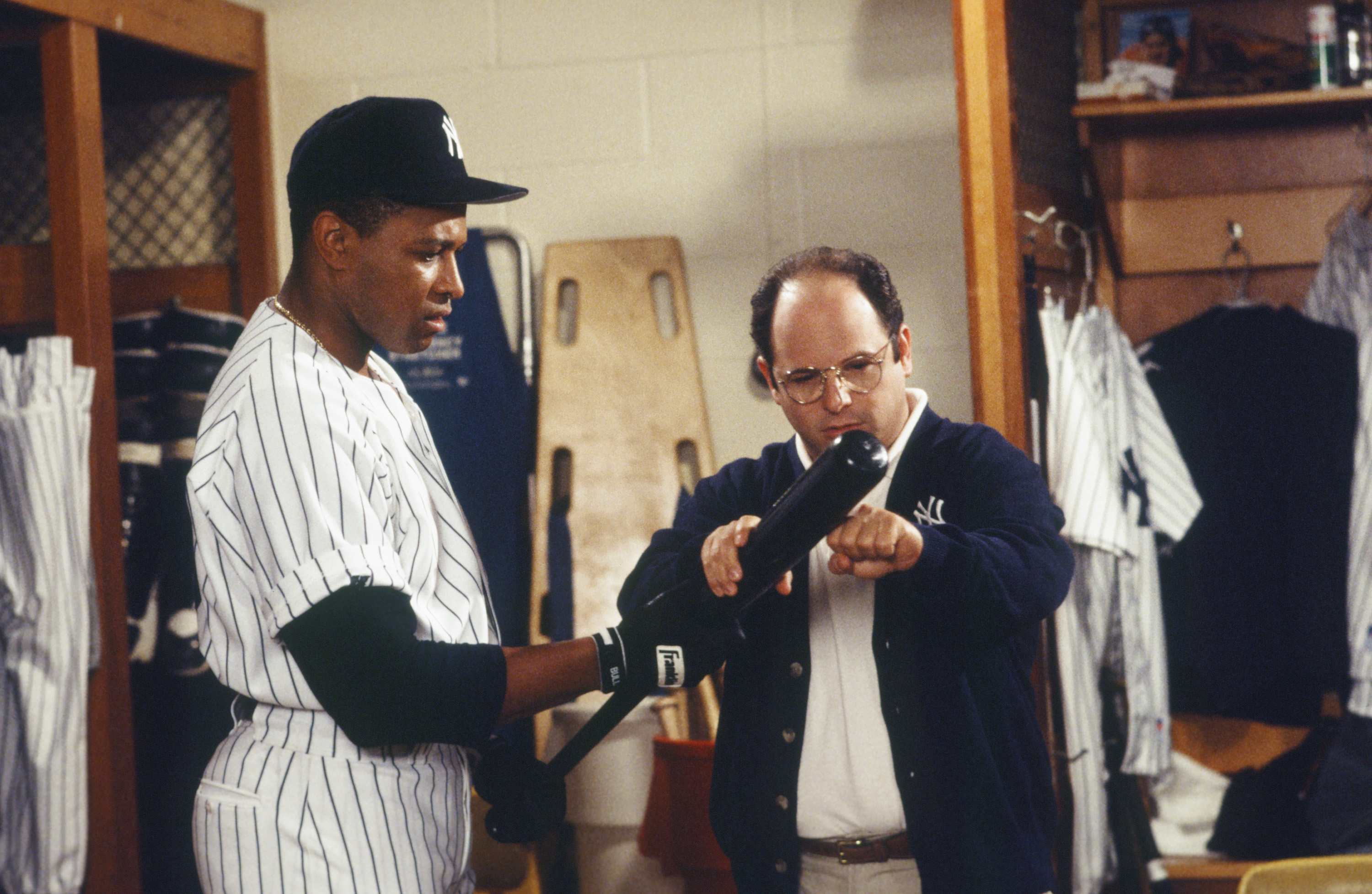 A baseball player in a Yankees uniform is shown a bat by a man in glasses and a jacket in a locker room setting.