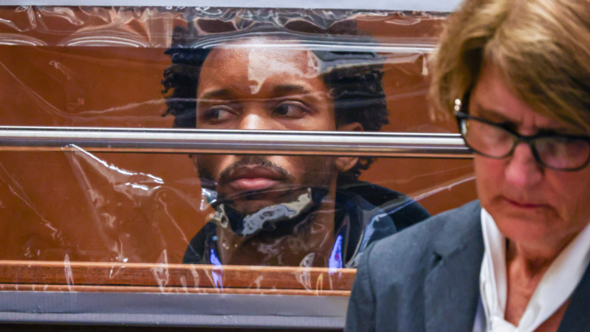 d4vd is seen through a transparent barrier, looking intently. A woman with glasses is in the foreground, focused on something off-camera.