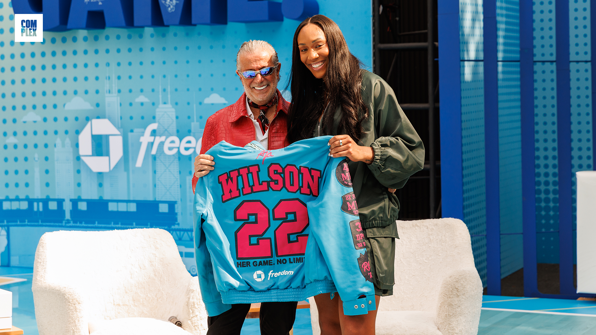 Two people smiling and holding a blue jacket with "WILSON 22" on it. The setting is a stage with blue decor and white chairs.