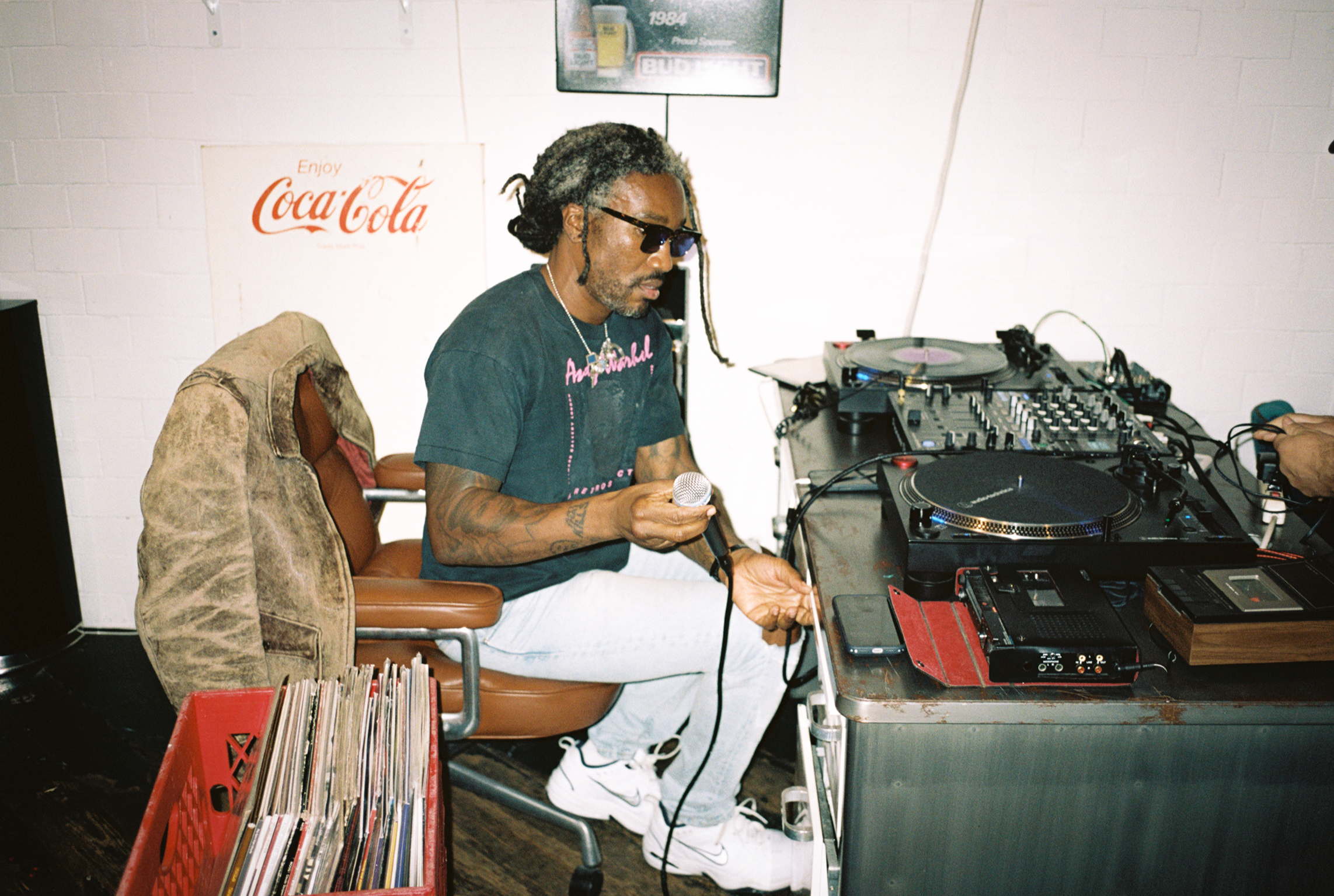 A person with sunglasses holding a microphone sits by DJ equipment, surrounded by vinyl records and a Coca-Cola sign in the background.