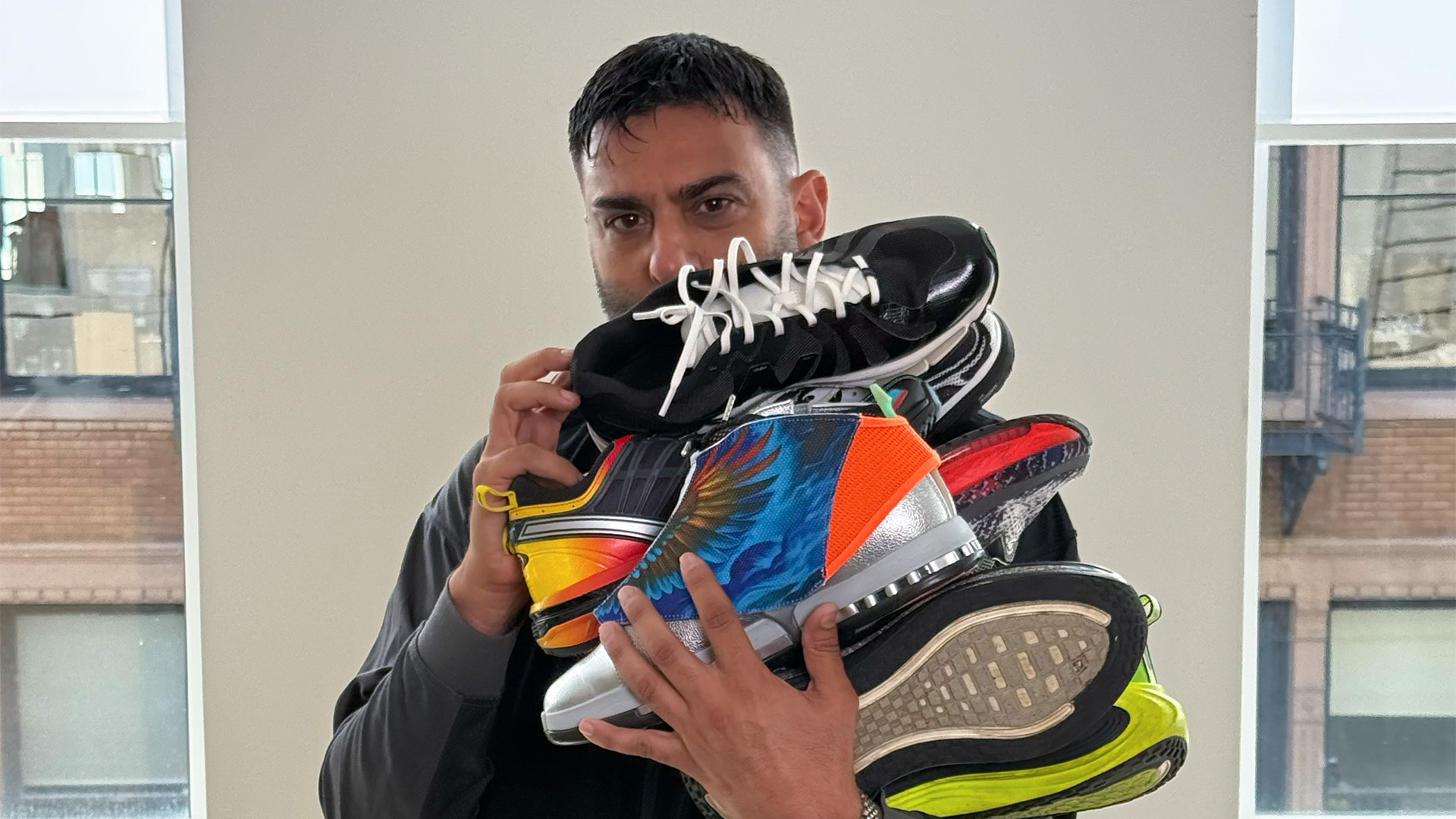 A person holding a stack of colorful sneakers in front of their face, with a cityscape visible through the window behind them.