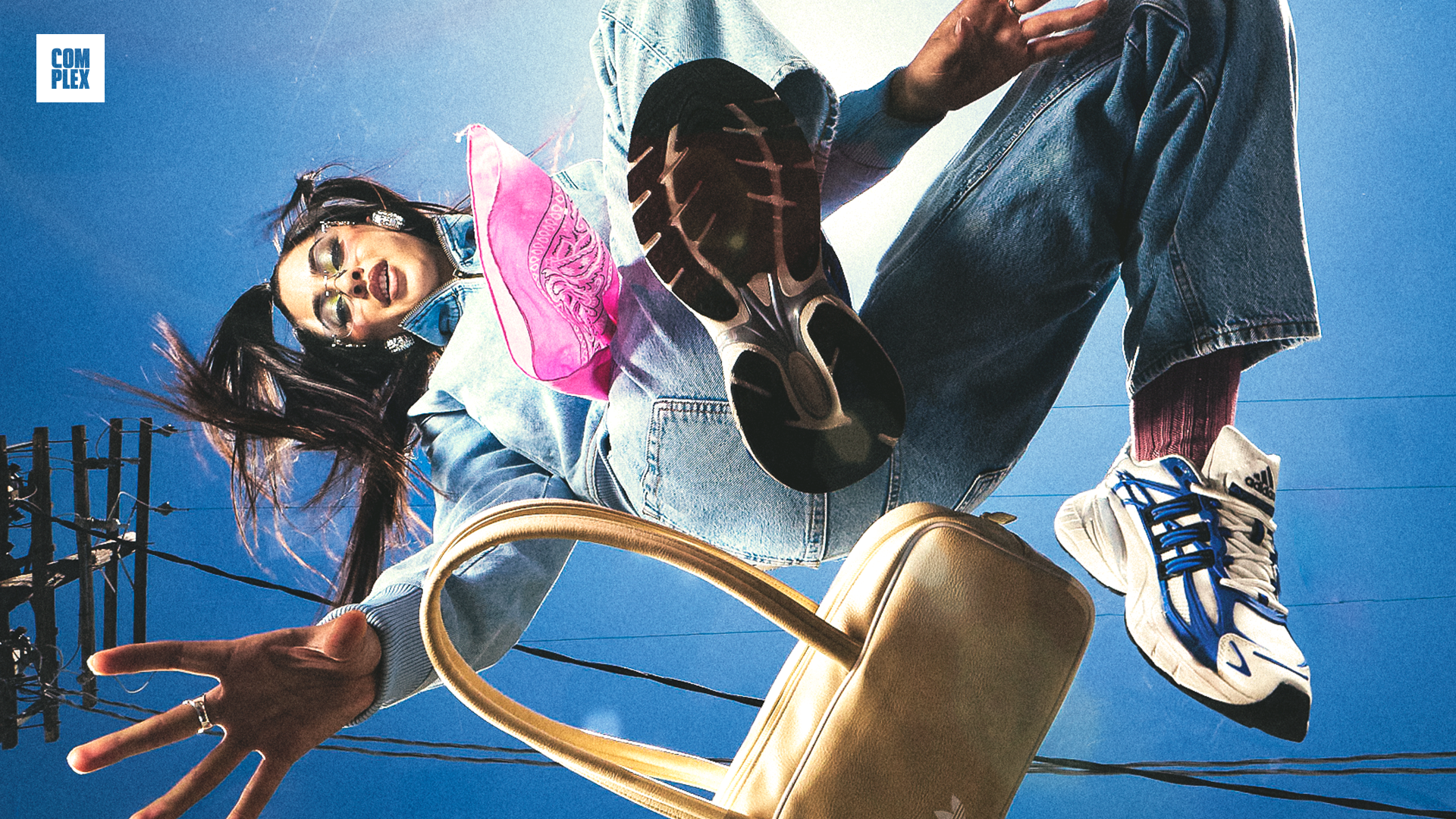 A person in mid-air wearing jeans, sneakers, a pink bag, and headphones, with a dramatic low-angle view against a blue sky.