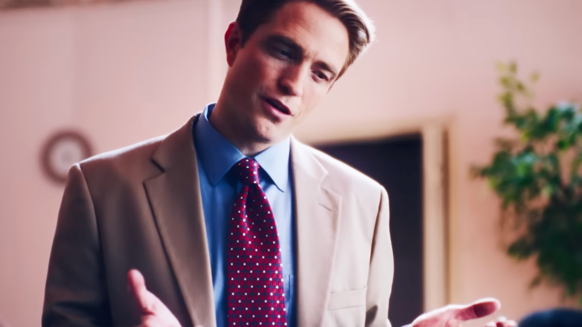 Robert Pattinson as Chris Hansen in a beige suit and red polka dot tie gestures while speaking in an indoor setting.