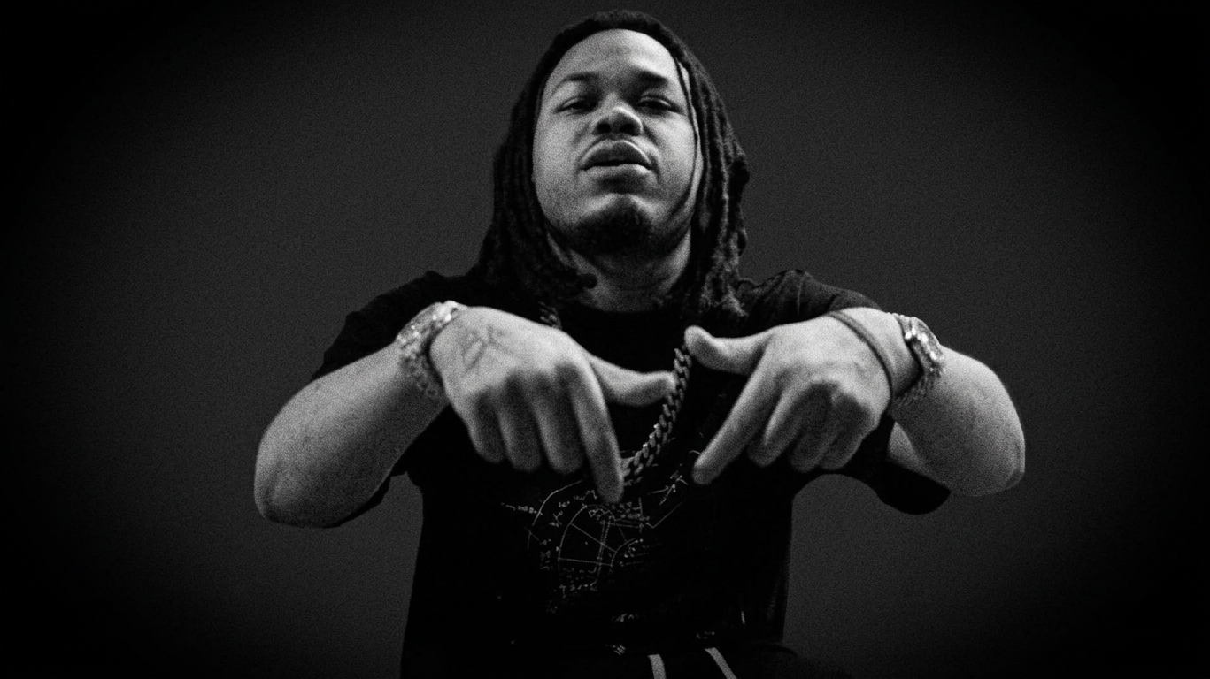 Bloodhound Q50 with dreadlocks, wearing a black shirt and jewelry, gestures with their hands against a dark background.