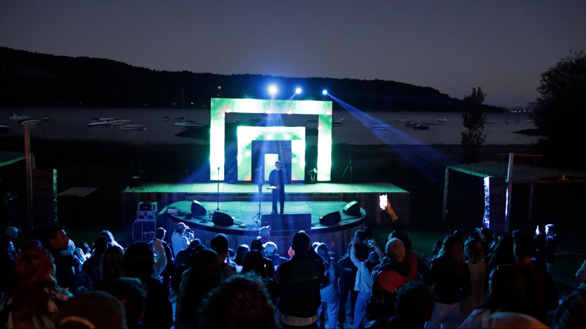 Outdoor concert at night with a lit stage and a performer. A crowd watches, with a lake and boats visible in the background.