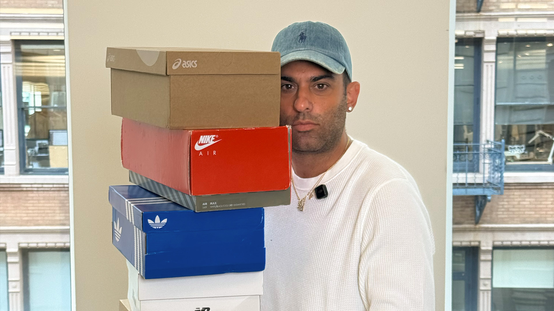 A person in a white shirt and blue cap holds a stack of shoeboxes from brands like Nike, Adidas, and Asics, against an urban window backdrop.