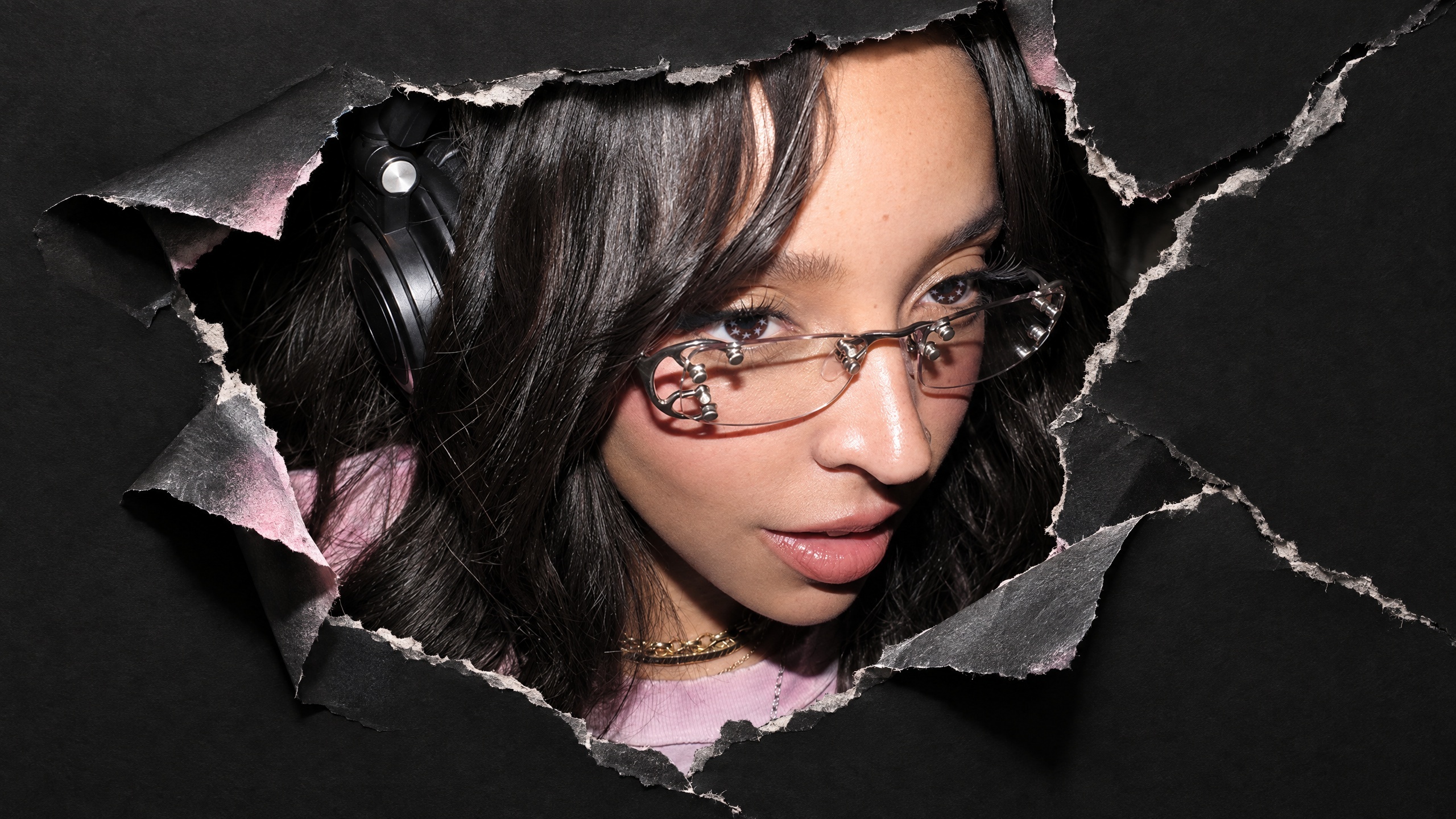Tinashe with headphones peers through a torn black paper background, wearing glasses and a pink top. This is the cover for her new album 'Popstar.'