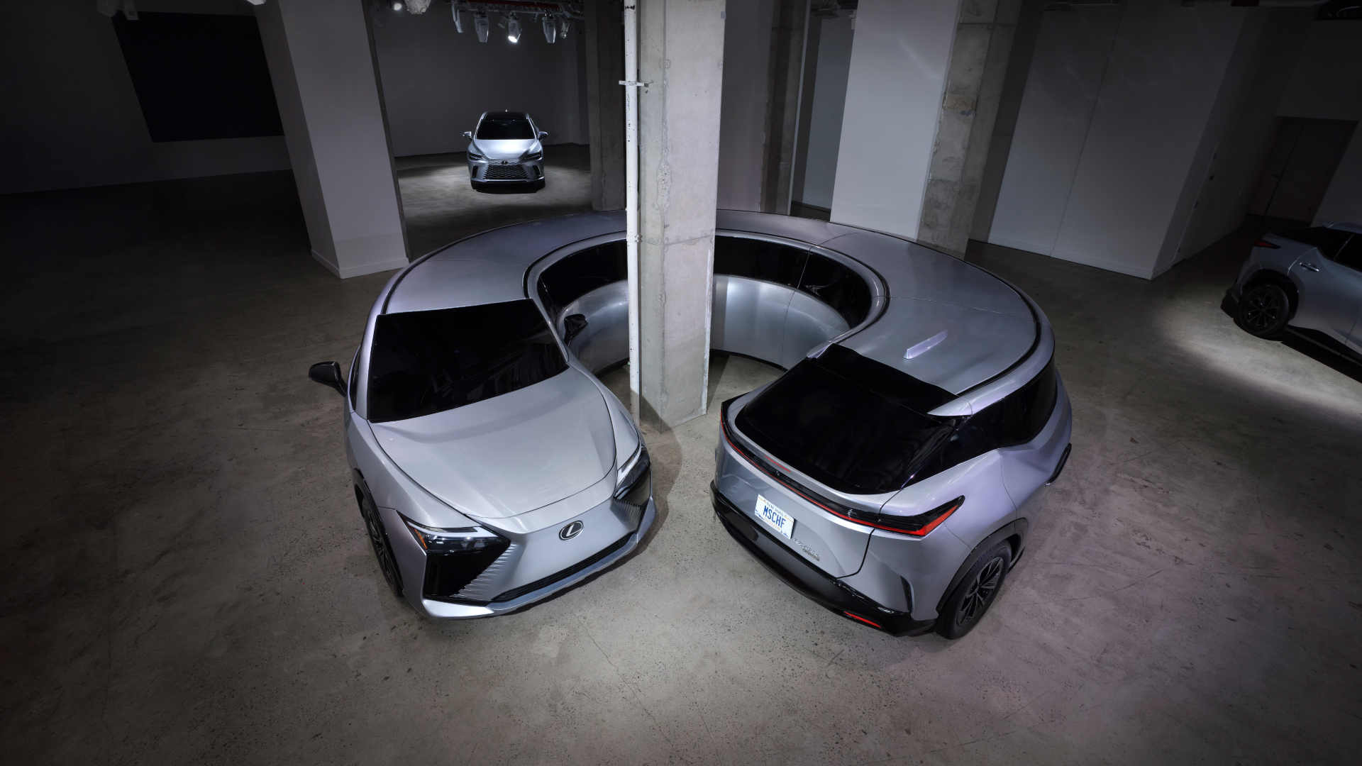 Two silver Lexus cars are parked in a showroom with a curved wall, showcasing their sleek design and modern features.
