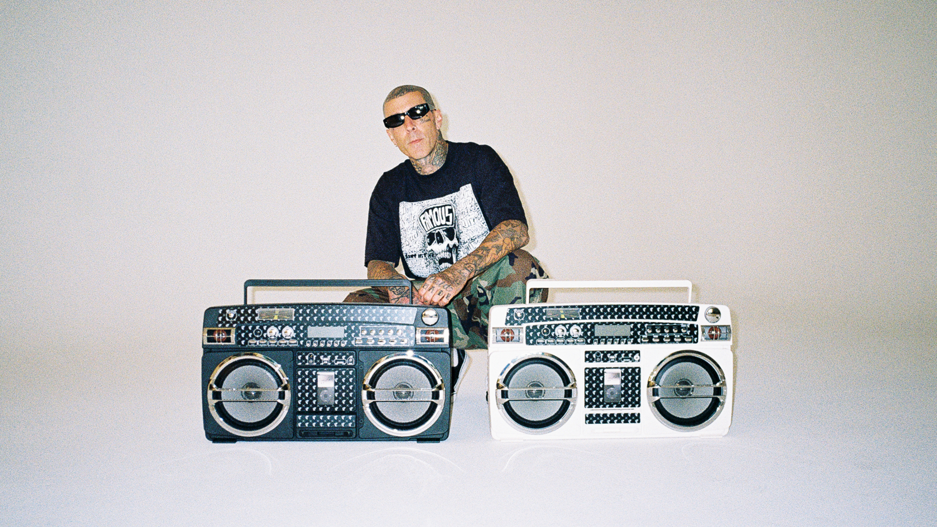 Travis Barker with tattoos and sunglasses sits behind two retro boomboxes, one black and one white, against a plain background.