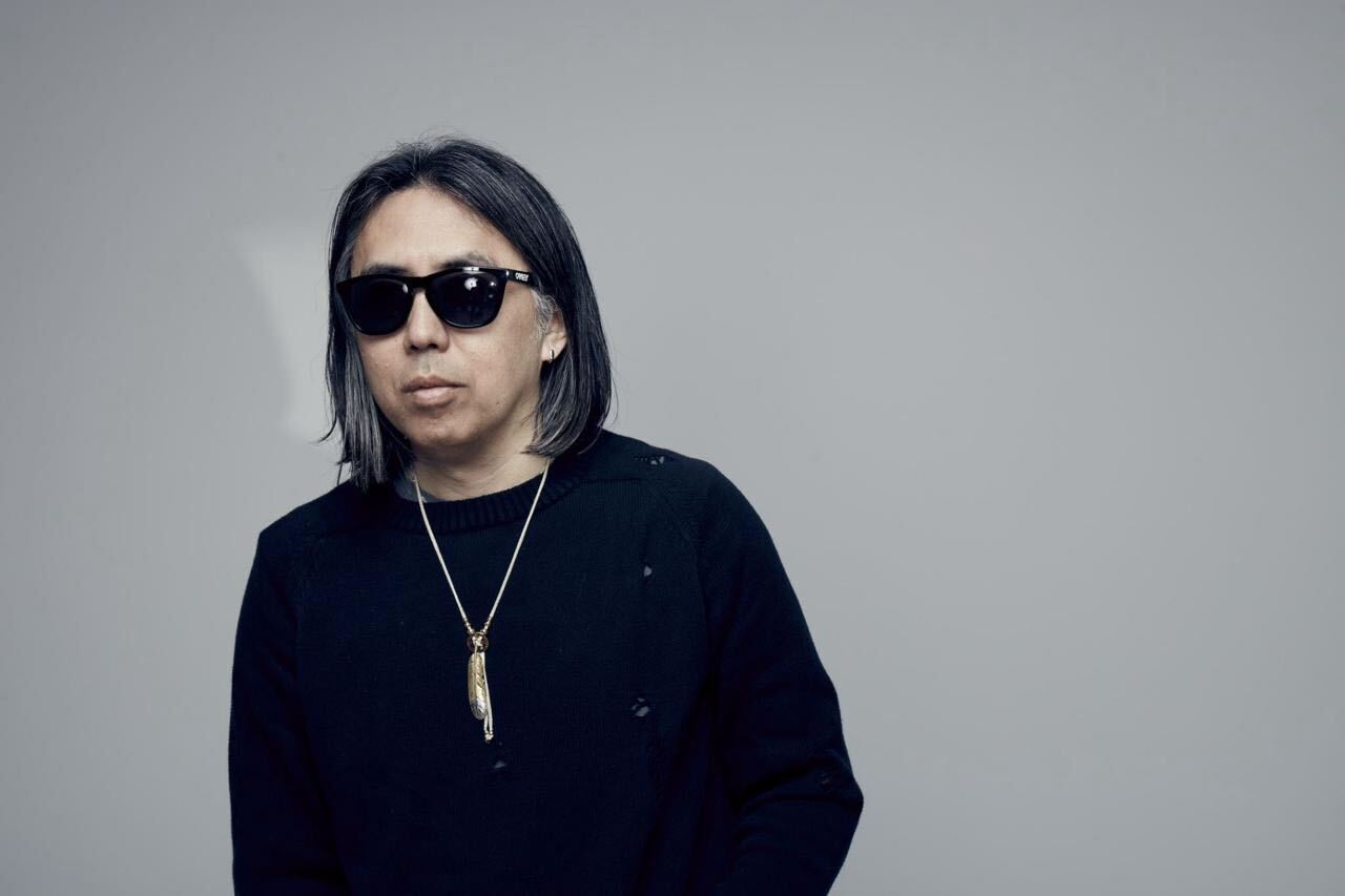 A person with shoulder-length hair, wearing sunglasses, a black shirt, and a necklace, stands against a plain background.