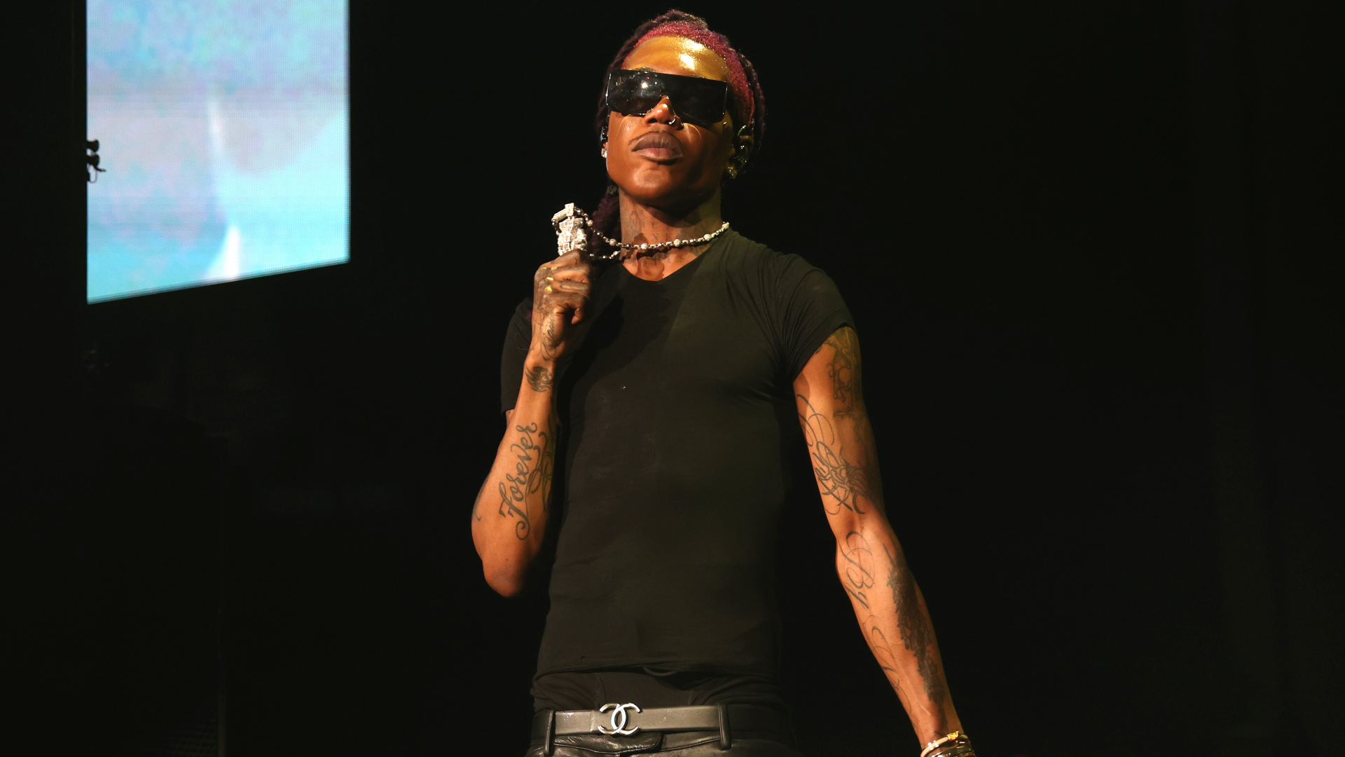 Destroy Lonely wearing sunglasses and a black t-shirt holds a microphone on stage.