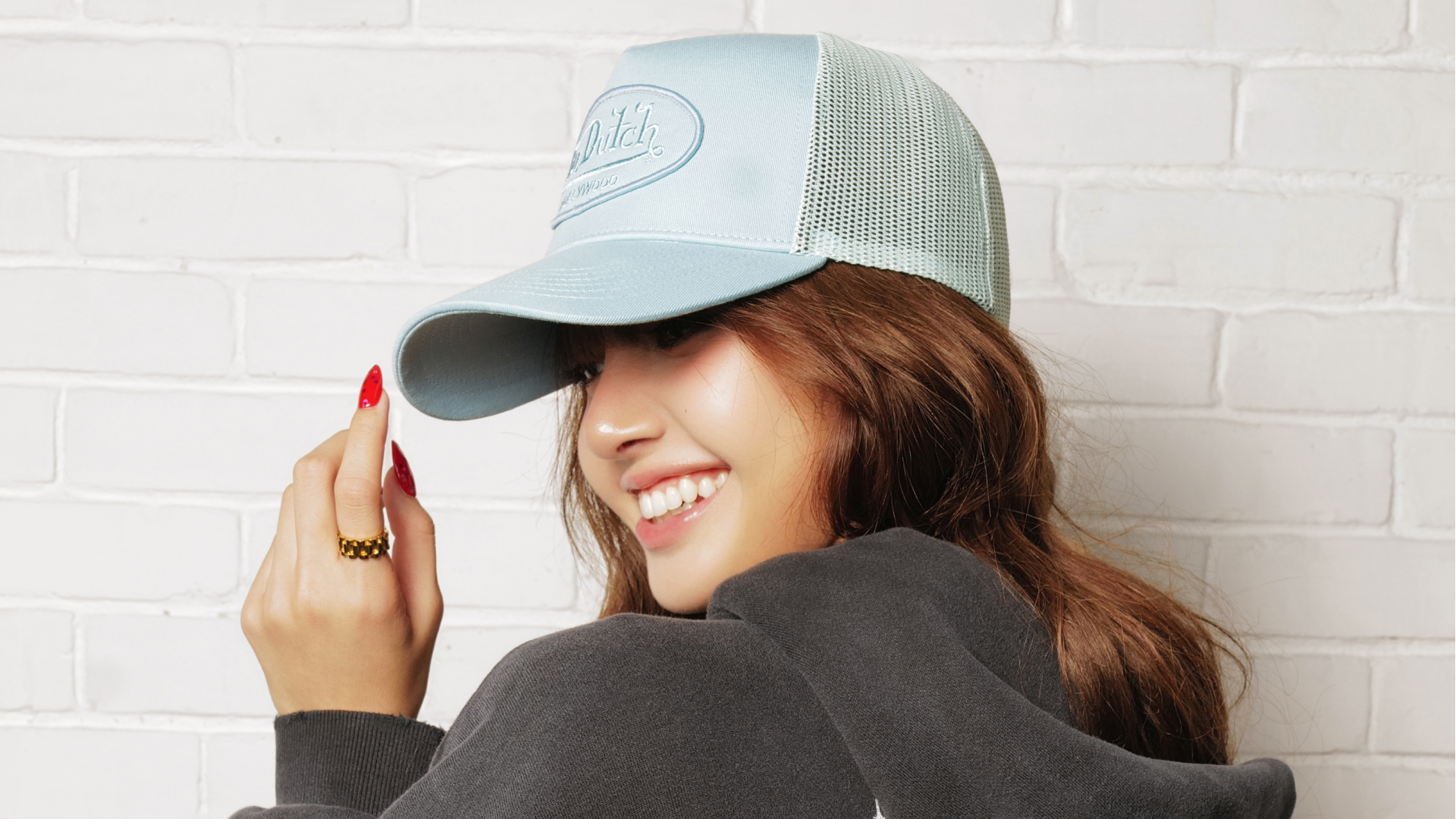 Woman in a light blue cap with red nails, smiling and looking to the side against a white brick wall.