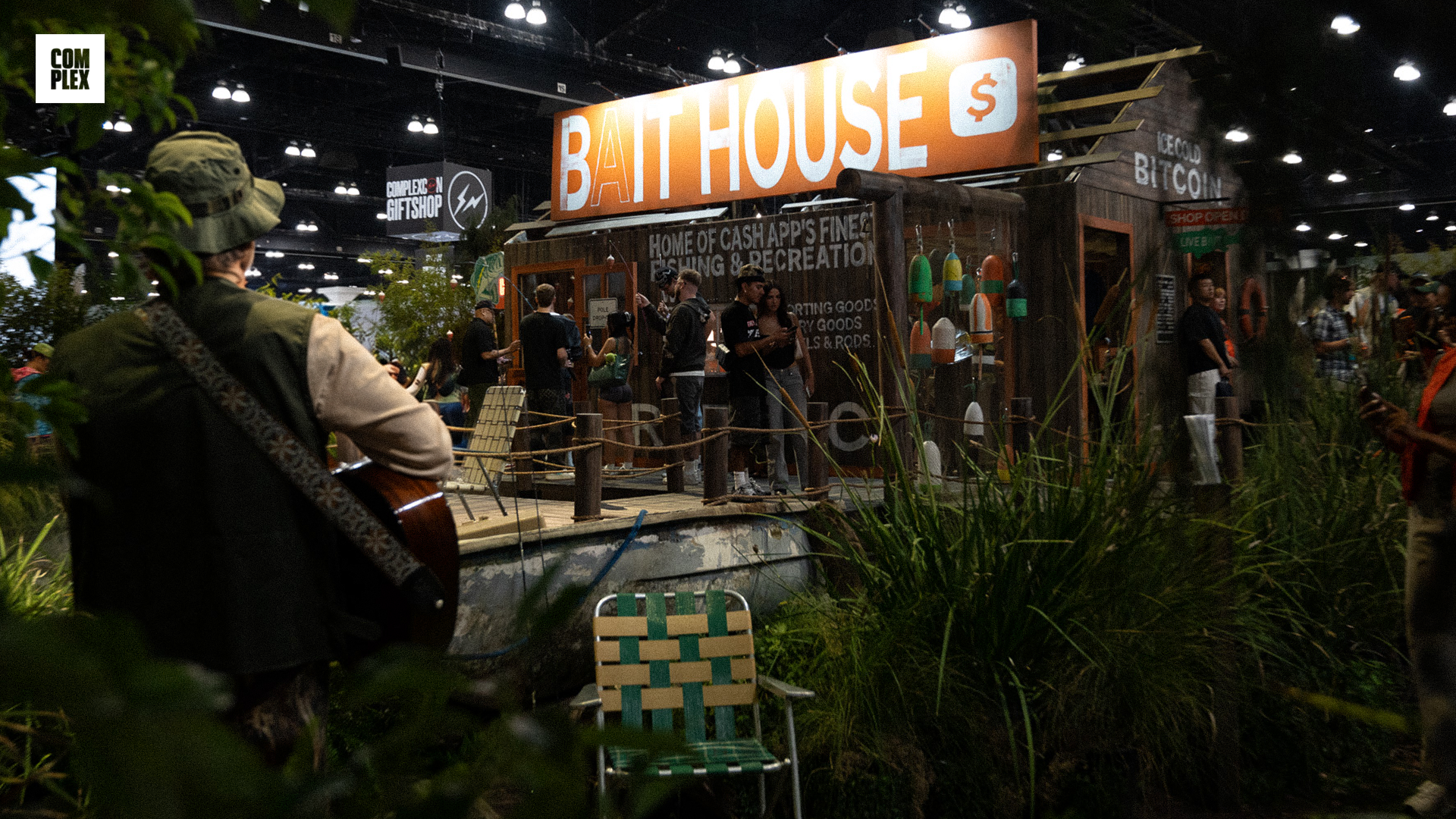 A "Bait House" booth at an event with people gathered around. A person with a guitar stands nearby, and greenery is in the foreground.