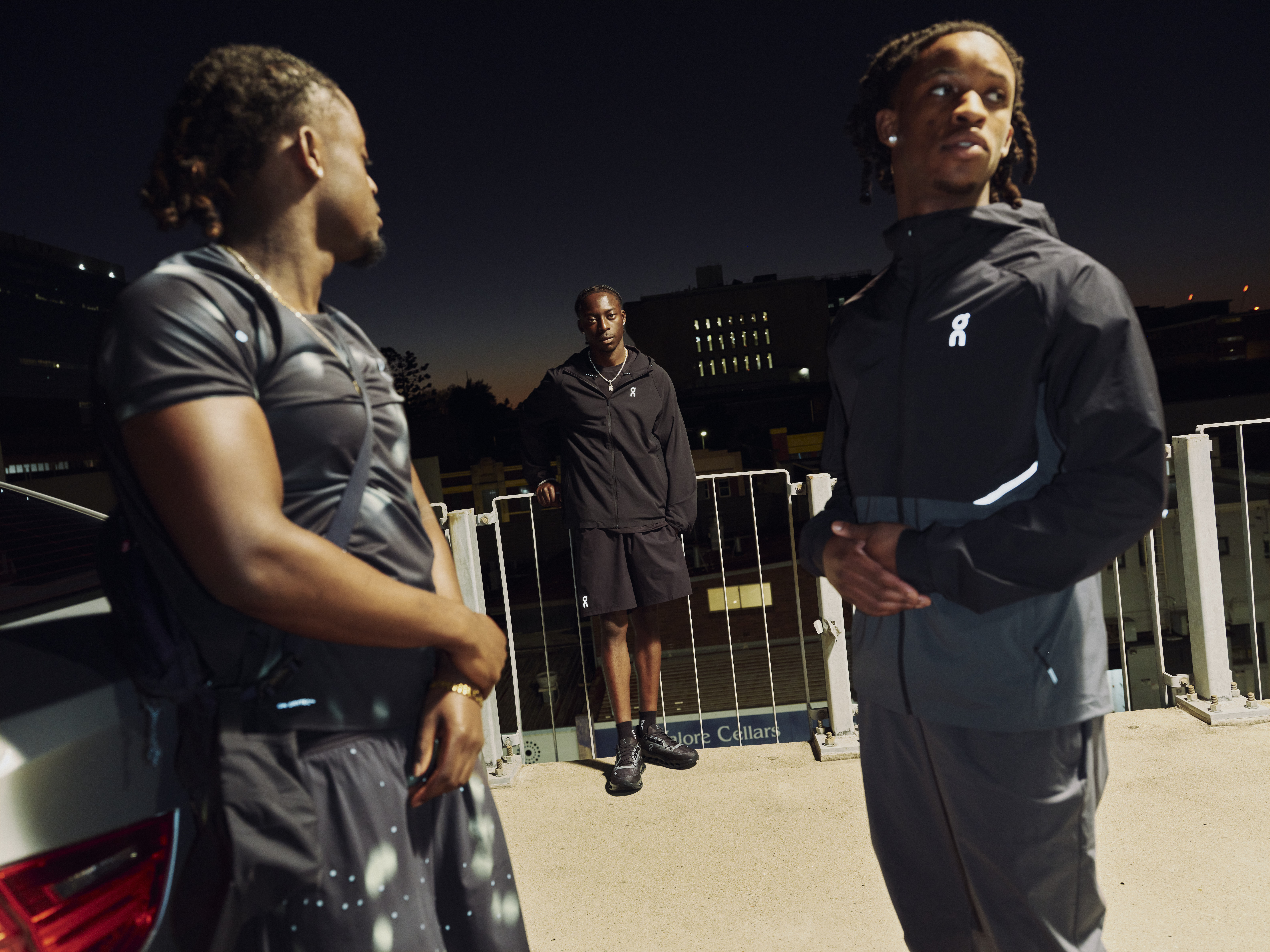 Three men in athletic wear stand on a rooftop at night, with a cityscape in the background.