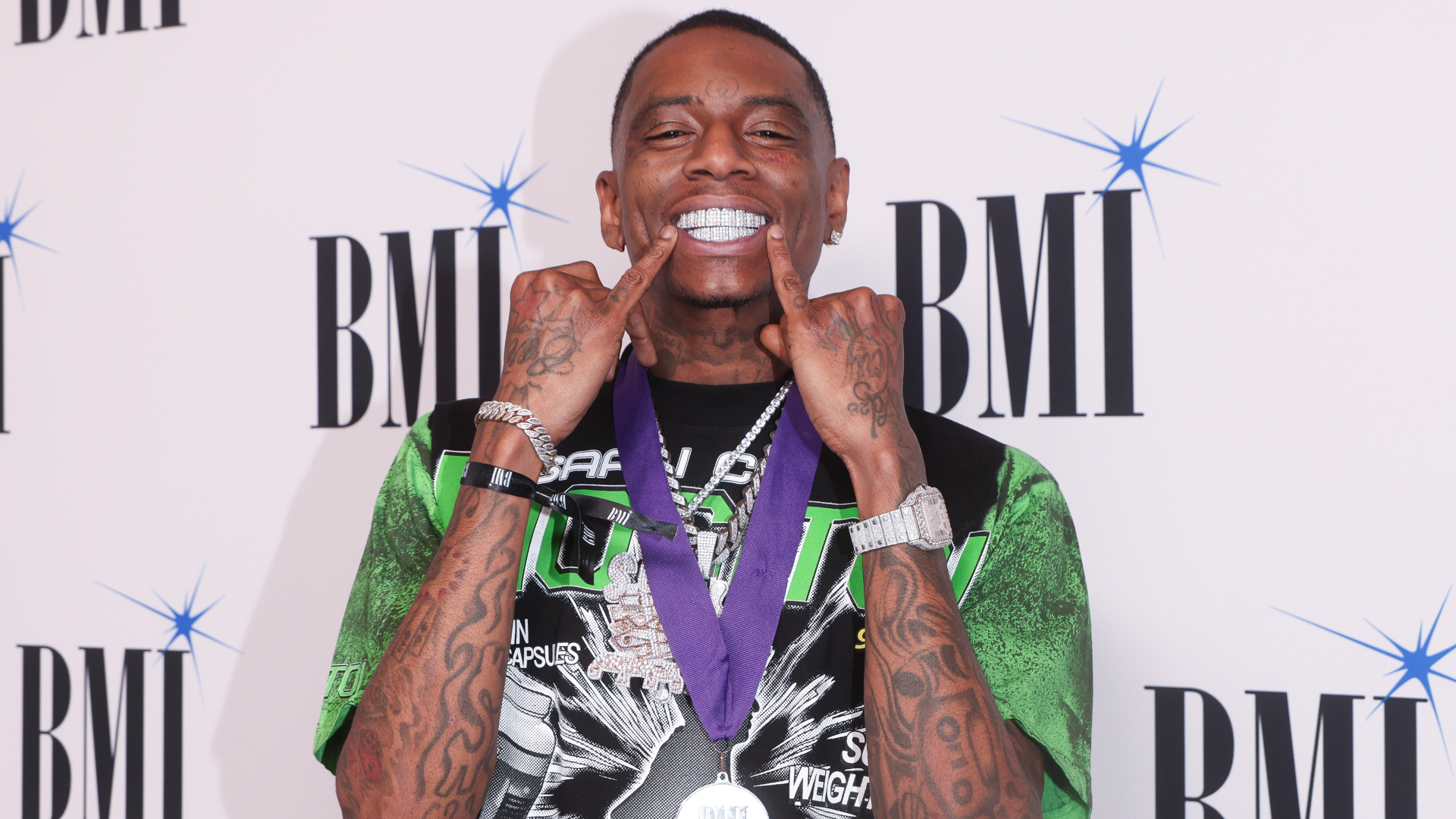 Soulja Boy smiling with diamond grills, wearing a green and black shirt, a purple lanyard, and jewelry, at a BMI event backdrop.