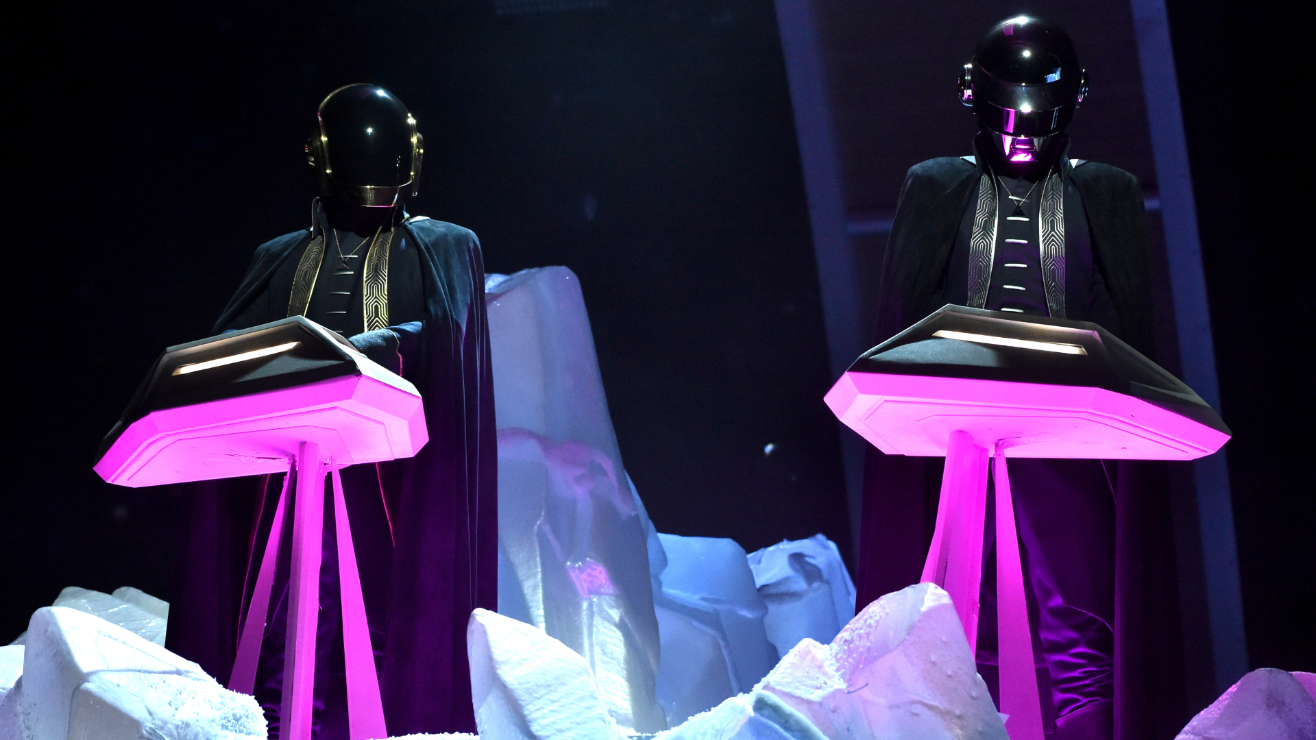 Daft Punk performing on stage, wearing their iconic helmets and black outfits, standing at futuristic consoles with purple lighting.