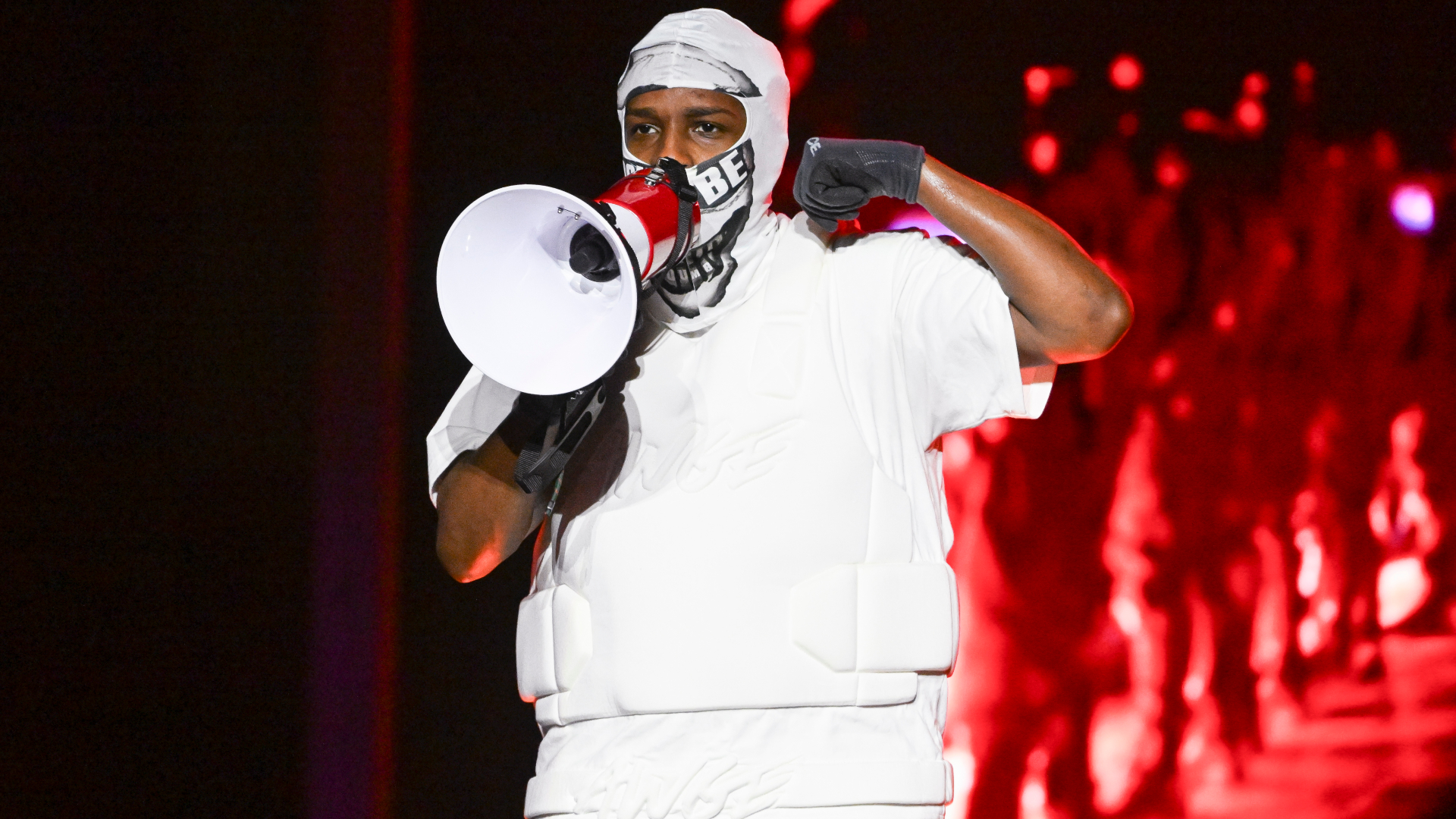 ASAP Rocky wearing a white outfit and mask, holding a megaphone on stage with a red-lit background.