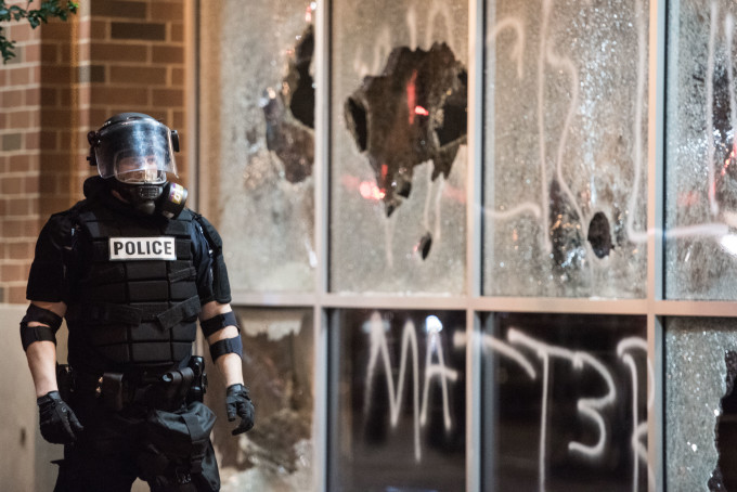 Police officer in riot gear in front of vandalized storefront.
