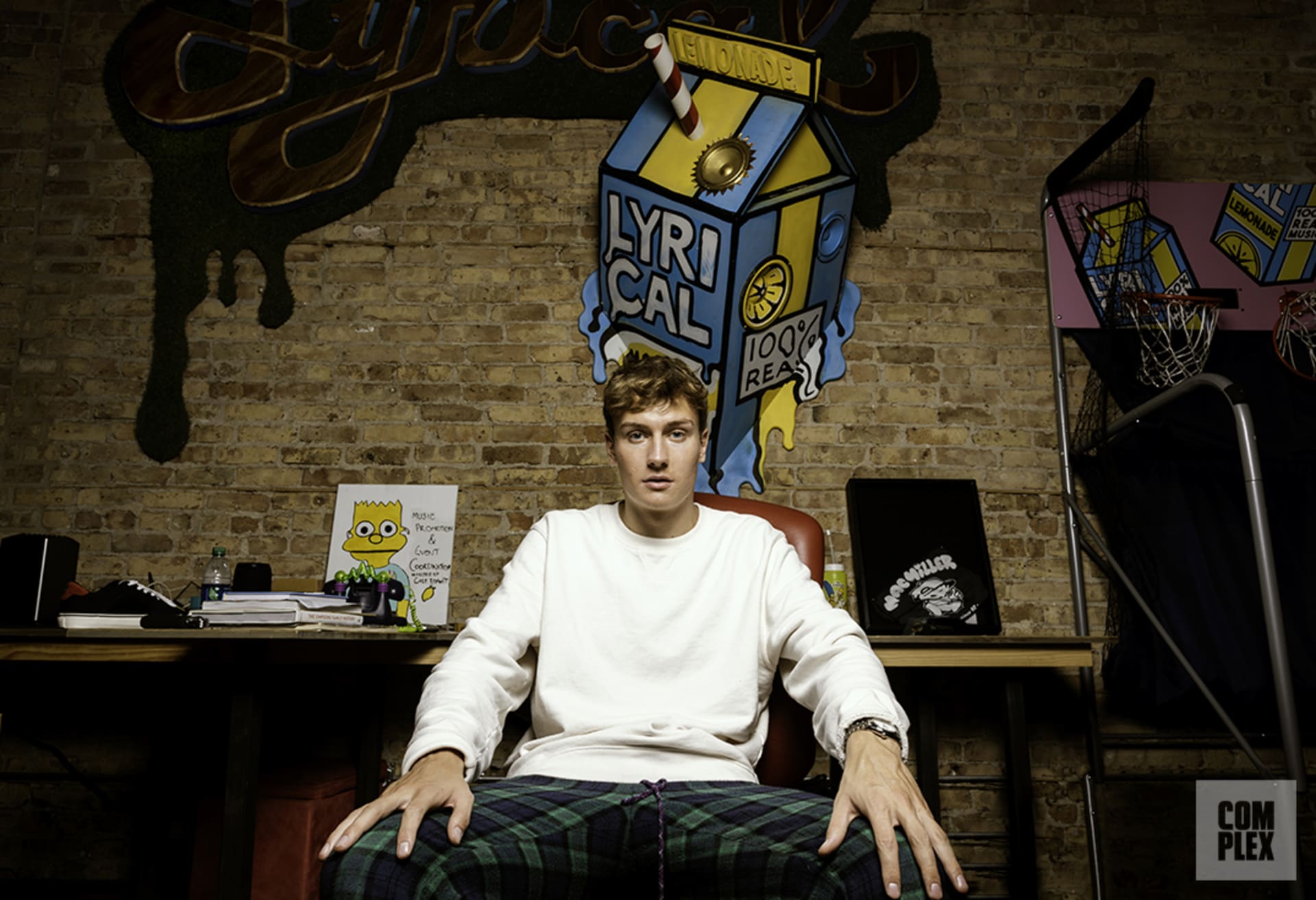 Photo of Cole Bennett, who is a white man with short blonde hair. He is wearing a long sleeve white shirt and green and navy plaid pants. Behind him, you can see a brick wall with a bunch of Lyrical Lemonade materials. Source: [link] https://www.complex.com/music/2019/07/cole-bennett-interview-lyrical-lemonade-chicago

