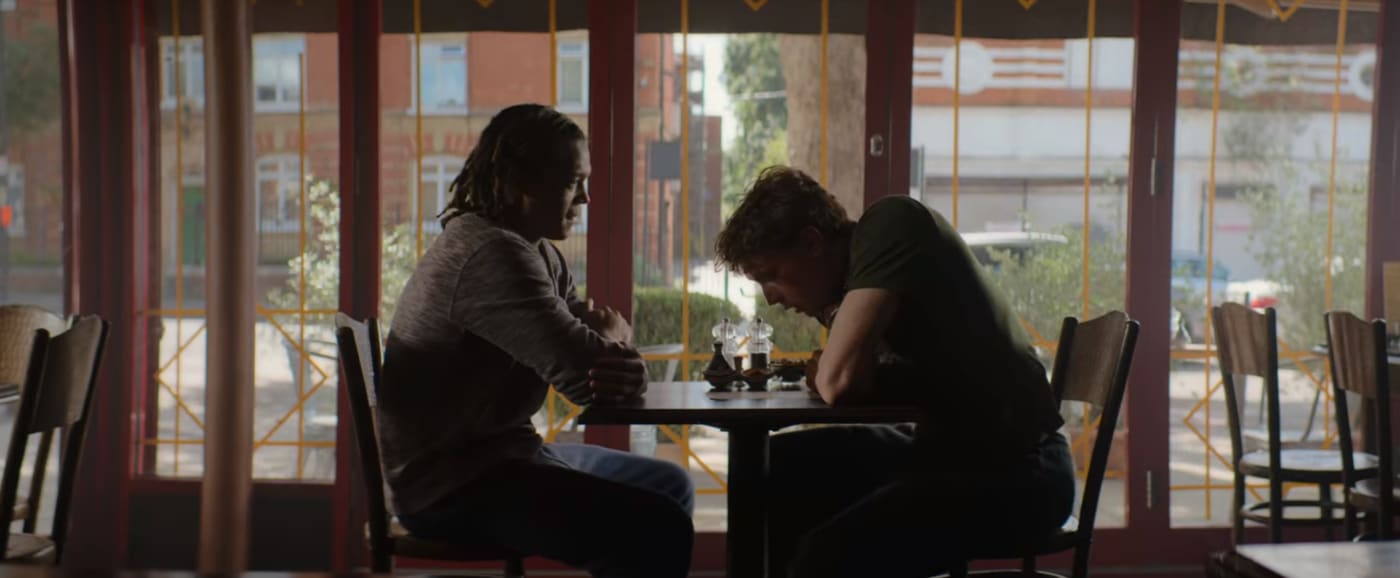 Two characters sit at a table and talk.