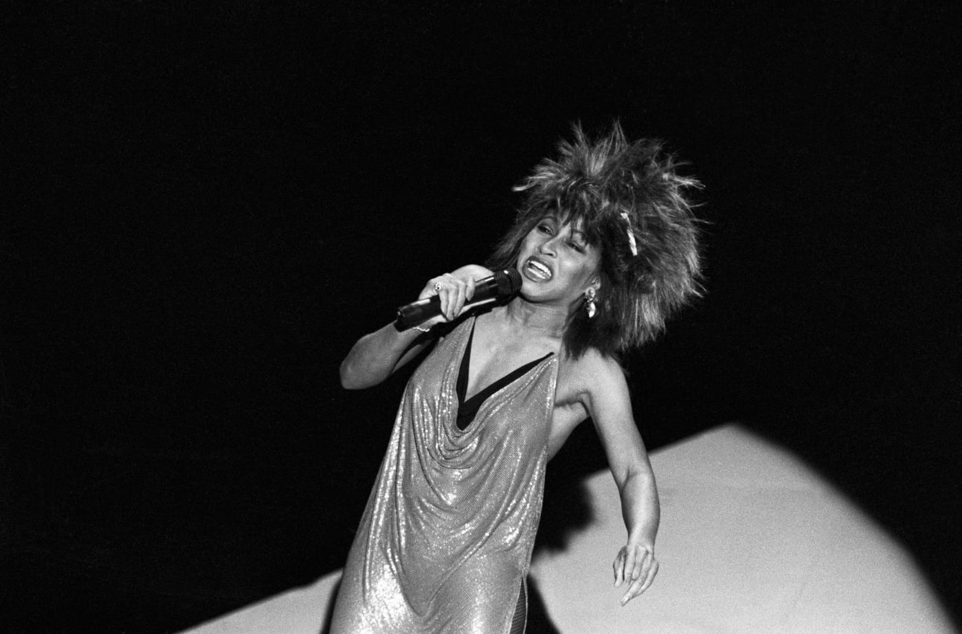 Black and white photo of Tina Turner in shiny dress passionately singing into microphone