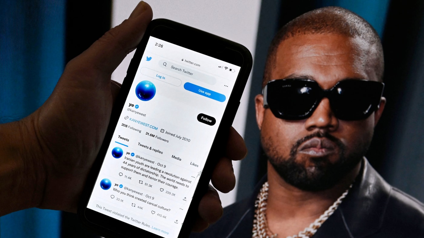 Twitter account of Kanye West is displayed on a mobile phone with a photo of him shown in the background.