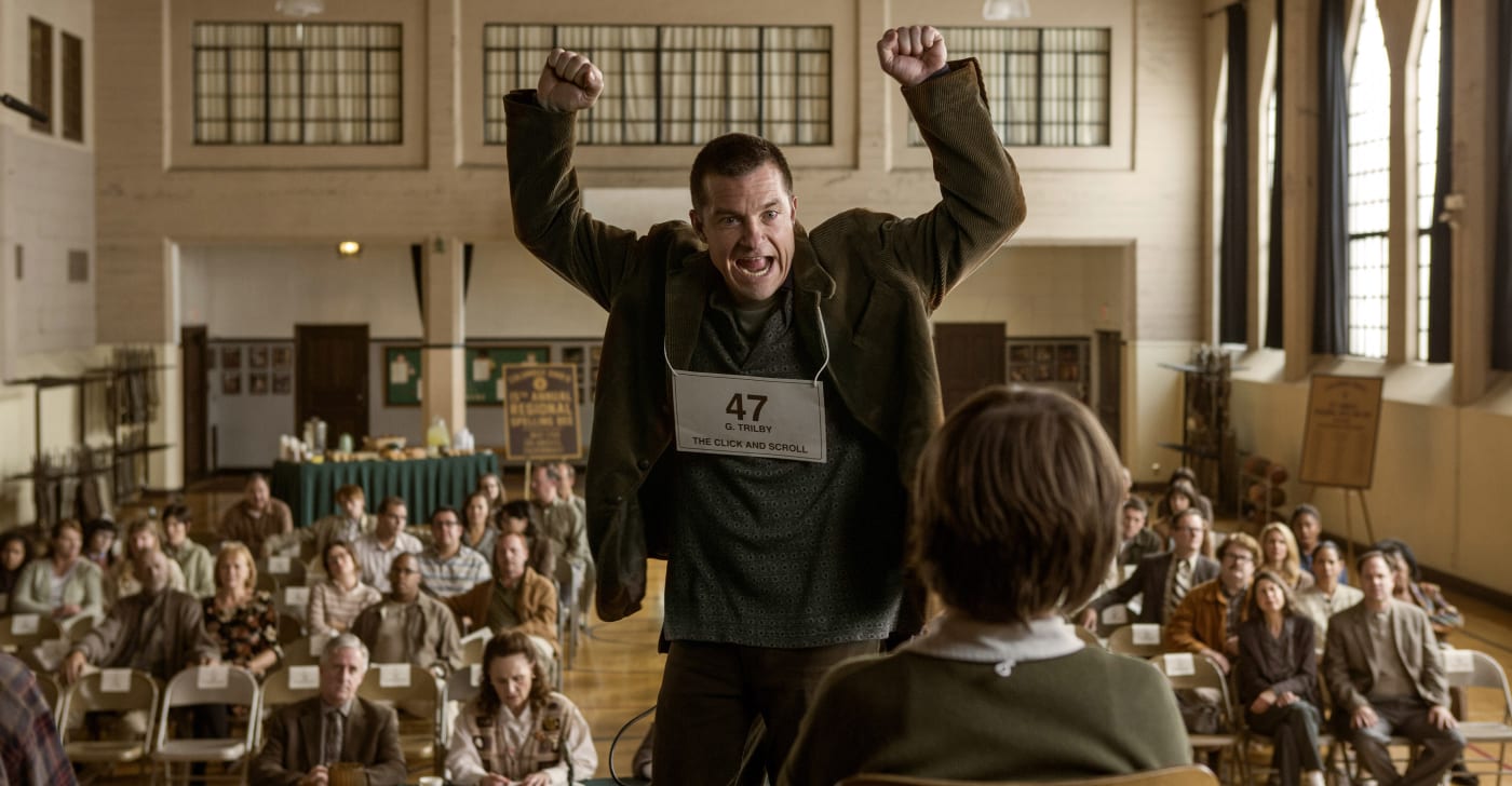 Jason Bateman raises both of his arms in the air.