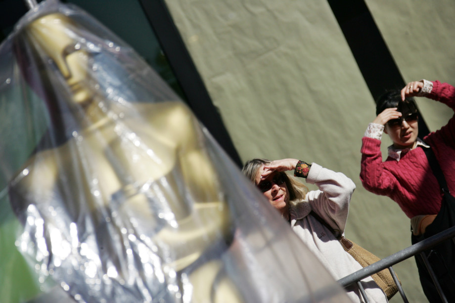 Two women stand next to an Oscar as they watch the Oscar Statues parade